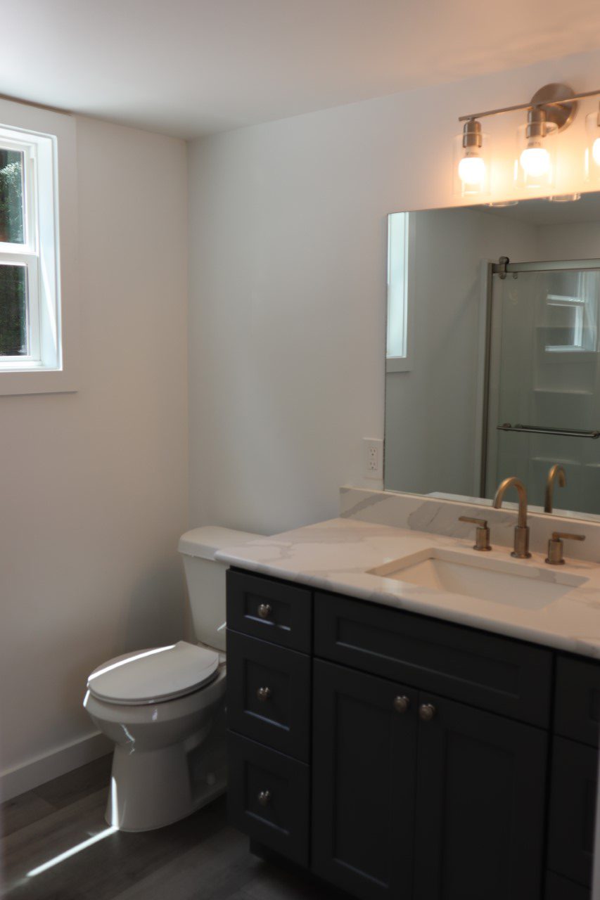 Modern bathroom featuring a dark vanity with brass fixtures, marble countertop, and large mirror. Natural light from a window adds a fresh feel.