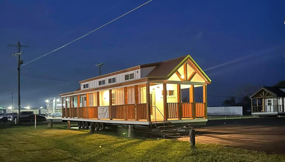 A warmly lit tiny house on wheels sits in a lot at dusk, its wooden exterior glowing amid dim surroundings. The scene feels cozy and inviting.
