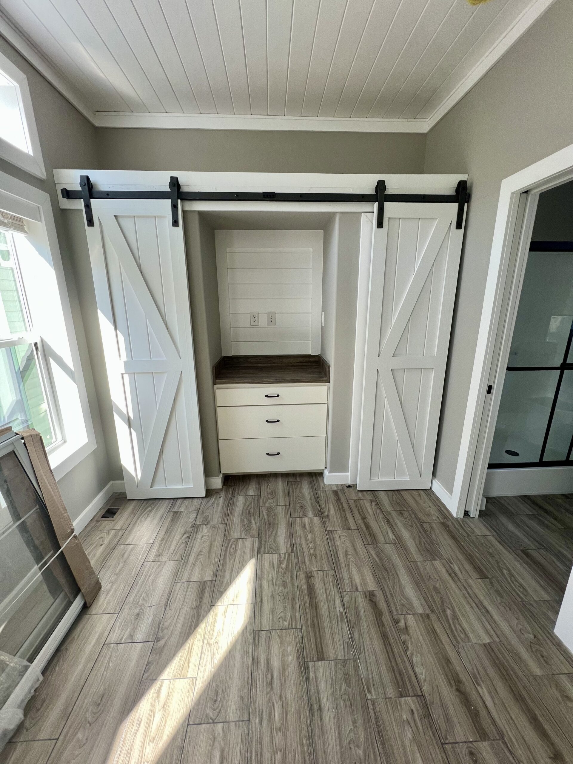 Bright room with wood-patterned tile flooring and sliding barn doors, revealing a small alcove with three drawers and a countertop. Natural light streams in from large windows, creating a calm and airy atmosphere.
