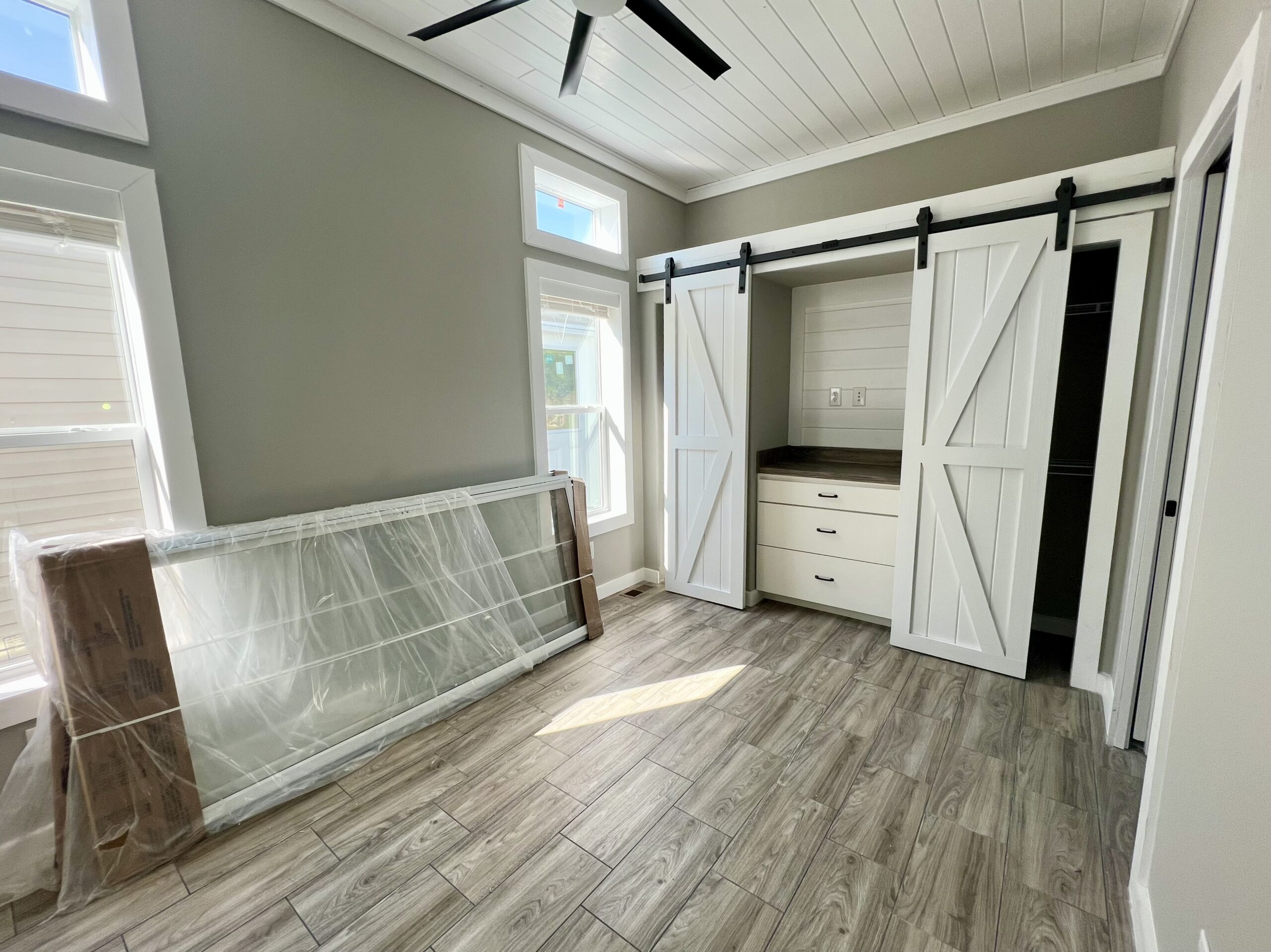 Bright room with wood-look tiled floor, gray walls, and white ceiling fan. Features a closet with white barn doors and a large window letting in sunlight.