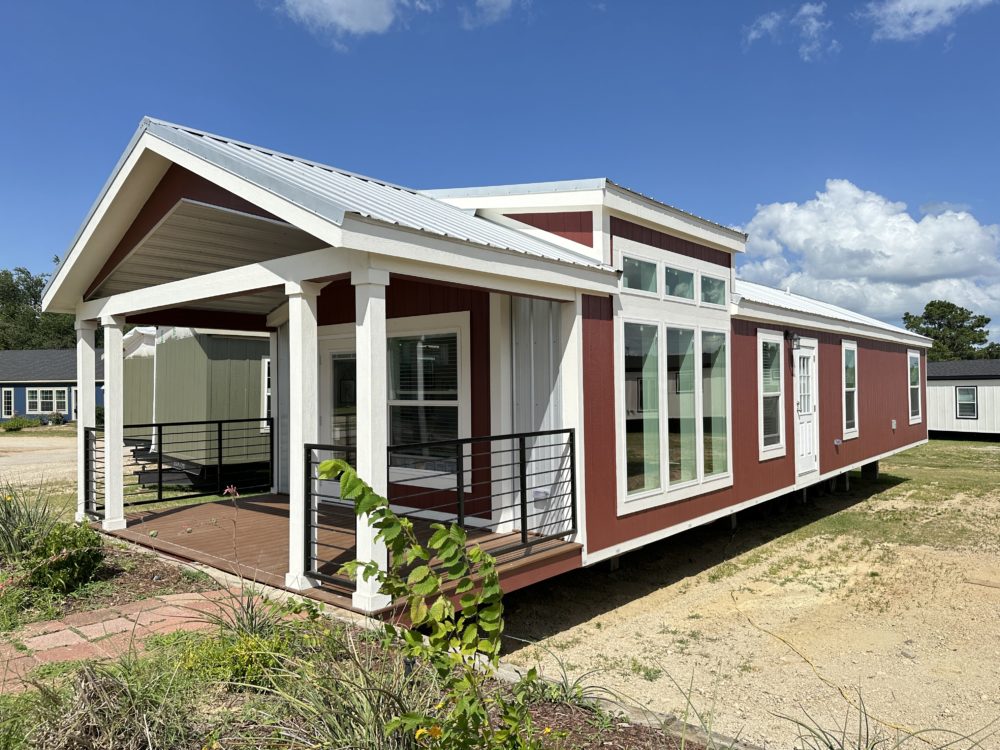 Red tiny house with white trim and metal roof, featuring large windows and a small porch with railing. Surrounded by grass and a few plants, under a partly cloudy sky.
