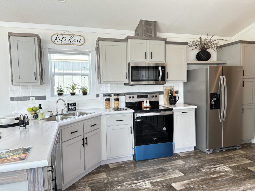 Modern kitchen with gray cabinets, stainless steel appliances, and wooden floors. Decor includes small plants on the windowsill, a fruit bowl, and a "Kitchen" sign. Bright and tidy atmosphere.