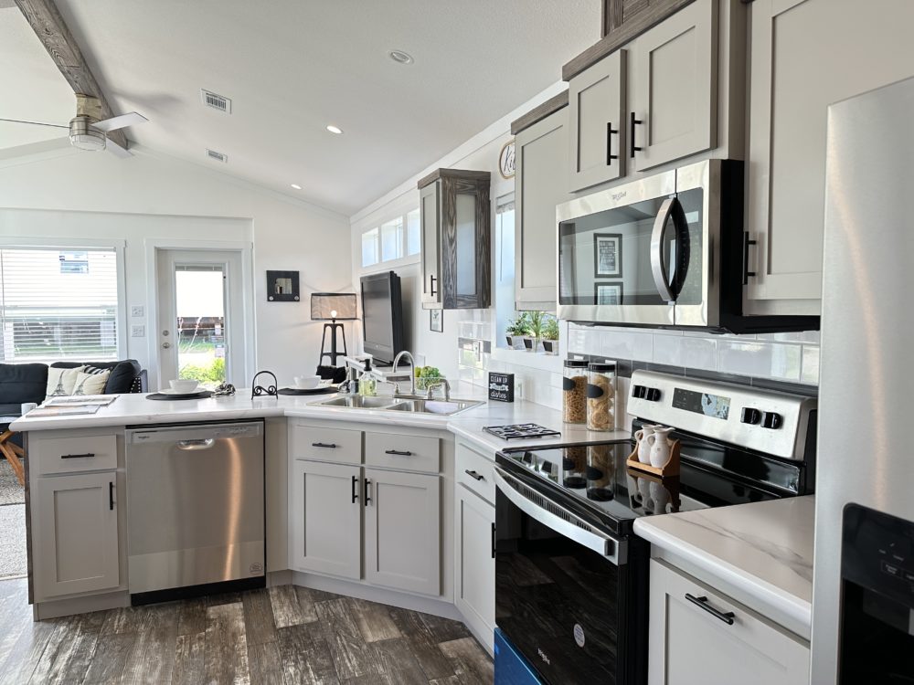 Modern kitchen with stainless steel appliances, gray cabinets, and wooden floor. Bright light from windows, with plants adding a fresh touch. Cozy and inviting atmosphere.