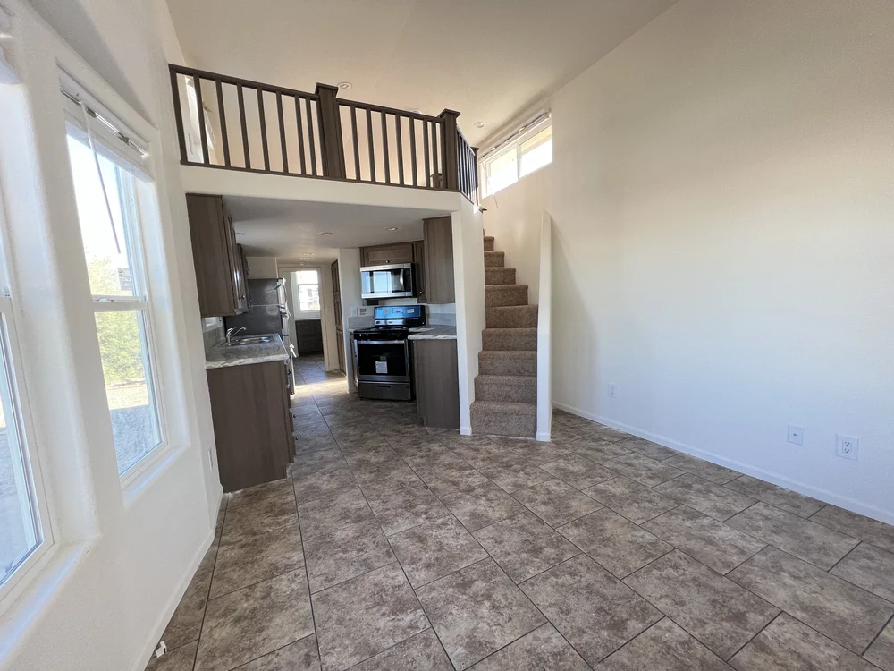 Modern, bright interior of a small home with tiled floors, brown kitchen cabinets, and a staircase leading to a loft. Large windows enhance spaciousness.