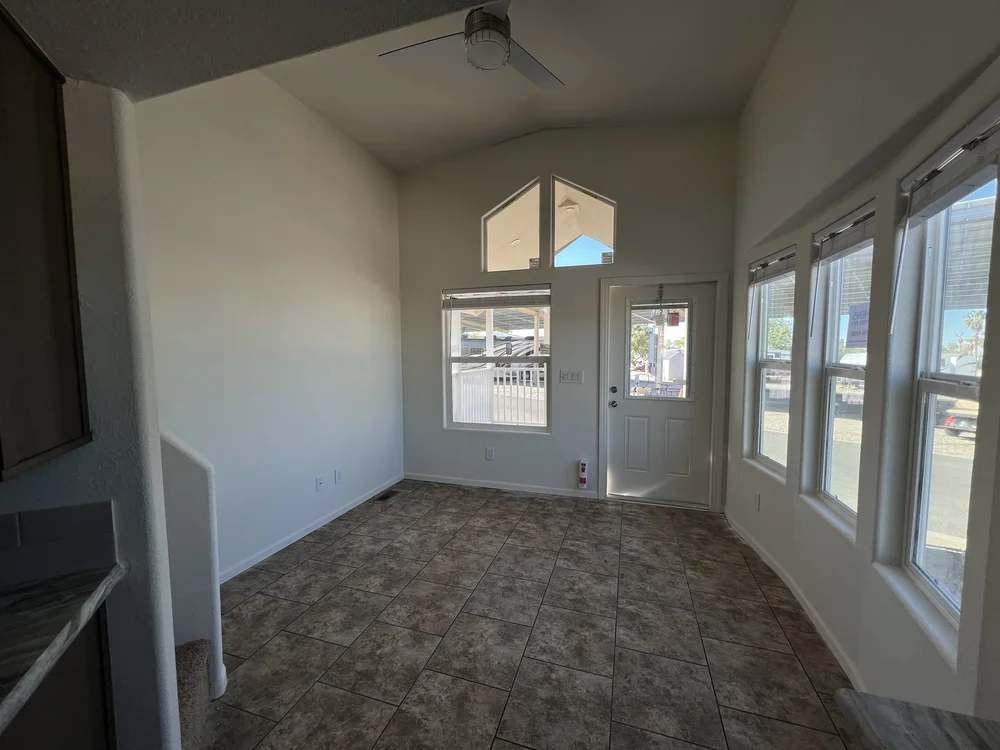 Bright room with high ceiling, pale walls, and tiled floor. Sunlight streams in through a white door and multiple windows, creating an airy feel.