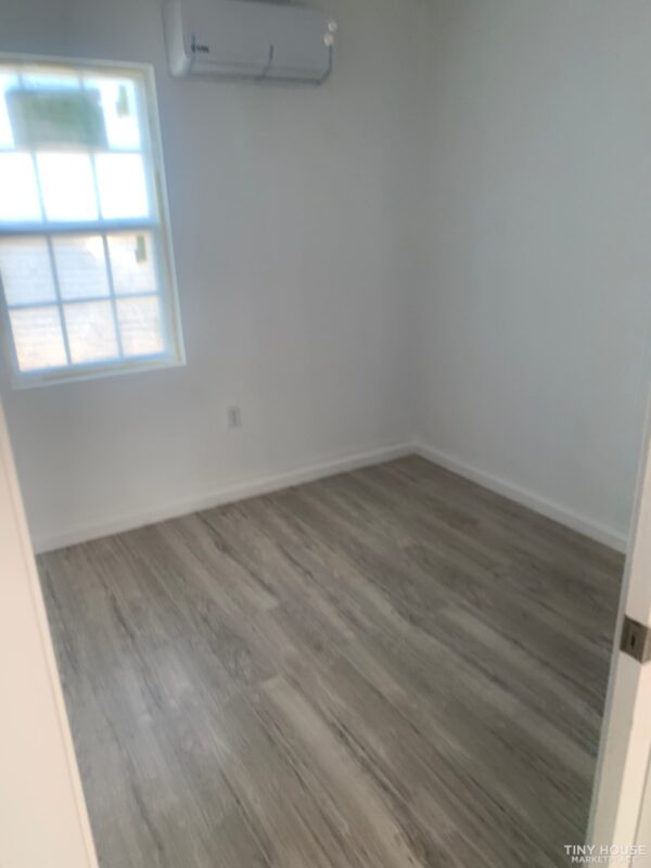 Small, empty room with light wood flooring and white walls, featuring a window with natural light and a wall-mounted air conditioner above. Minimalist and clean.