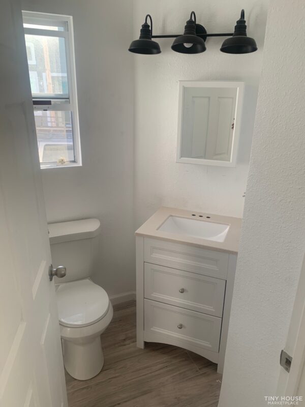 Small bathroom with a white vanity and sink, a mirror above, and a black triple-bulb light fixture. A toilet is beside a window, with wood-patterned floors.