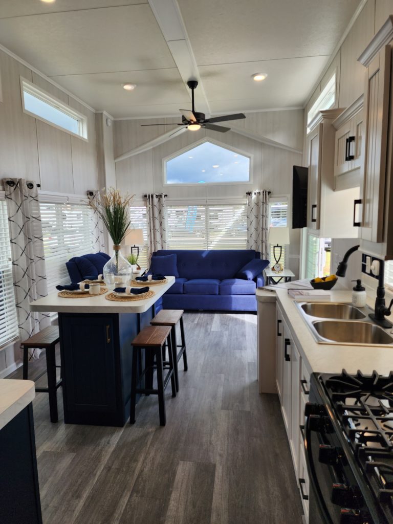 Modern kitchen and living area with light wood flooring and walls. Features a blue sofa, island with stools, and a ceiling fan. Bright, airy ambiance.