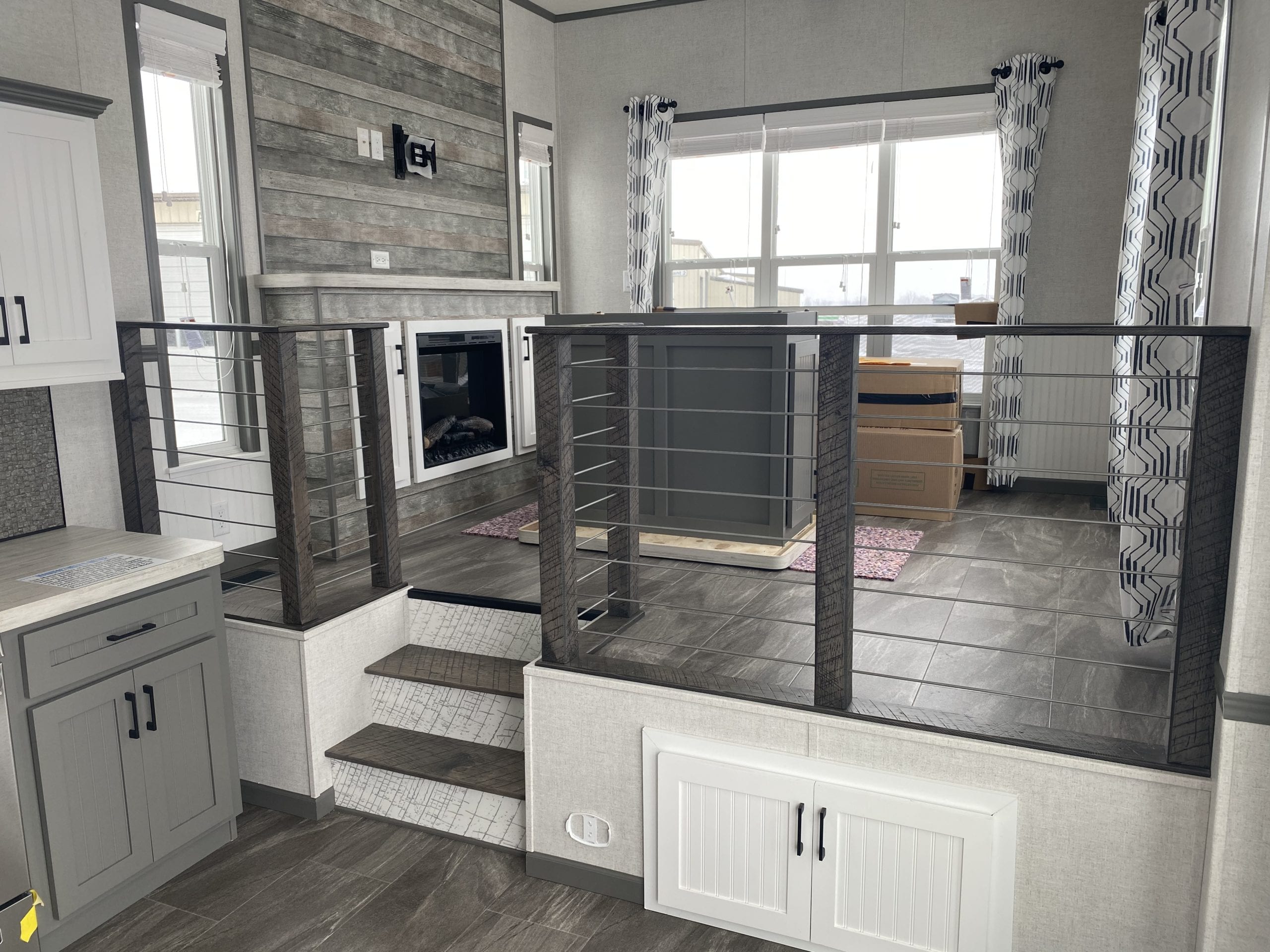 Modern living room with gray tones, featuring a fireplace, decorative railing, and large windows with patterned curtains. Cozy, contemporary feel.