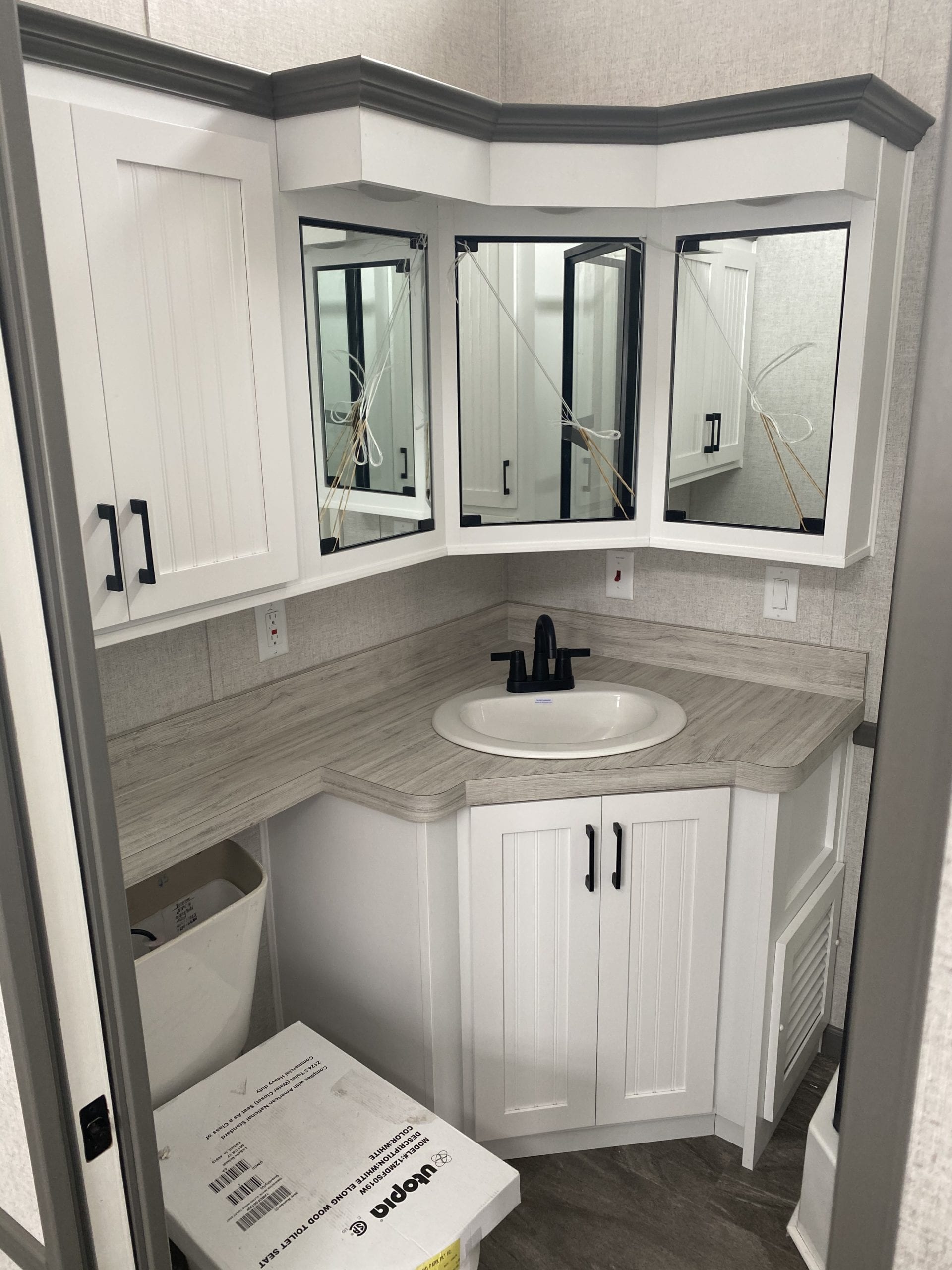 A bright, compact corner bathroom features a white countertop with an oval sink and black faucet. Above, mirrored cabinets are accented with dark trim.