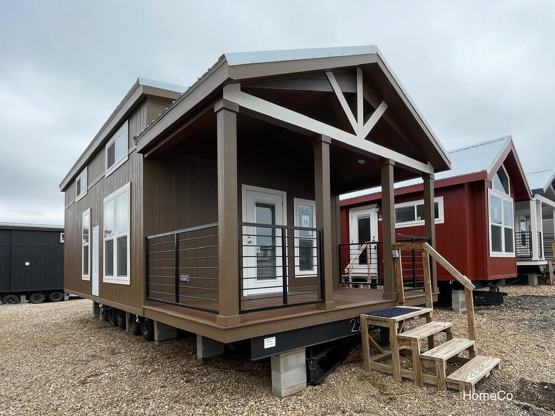 Compact brown tiny home with a front porch and railing, elevated on blocks. It features large windows and a pitched roof, with similar homes nearby.