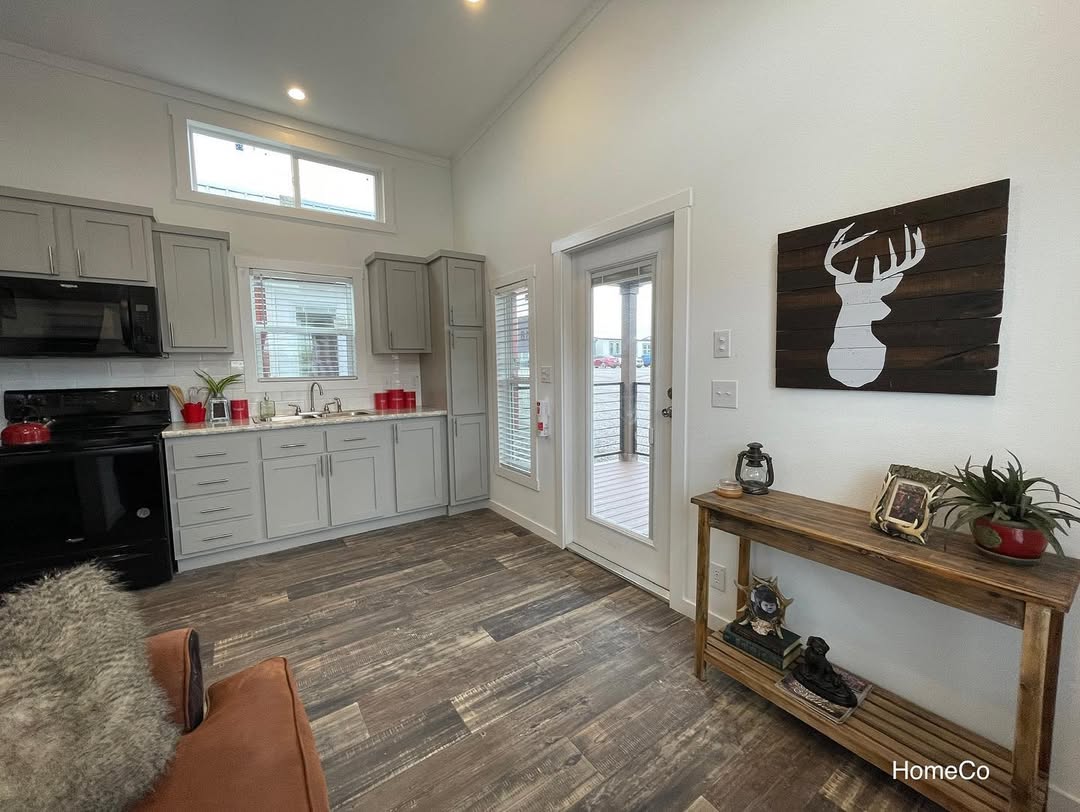 Modern kitchen-living room with gray cabinets, black appliances, and wood floors. Wall art of deer silhouette. Rustic table with decor items. Cozy ambiance.