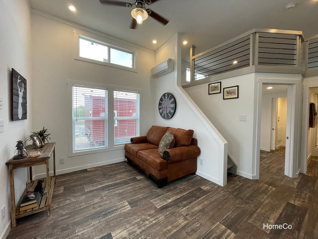 Cozy living room with a brown leather sofa, rustic wood floor, and modern decor. A staircase leads to an upper level, while soft lighting creates a warm ambiance.