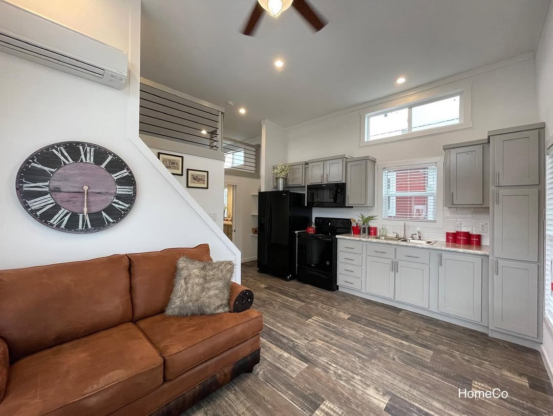 A cozy, modern living space featuring a brown leather couch with a gray pillow, a large wall clock, and a sleek kitchen with gray cabinets and black appliances.