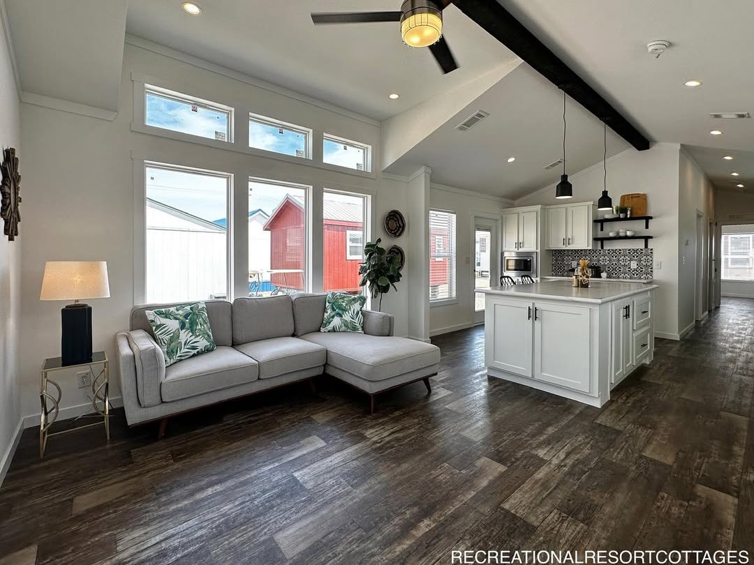 Modern living area with a light gray L-shaped sofa, tropical cushions, and a sleek floor lamp. Kitchen with white cabinets and island, wood flooring. Cozy and inviting.