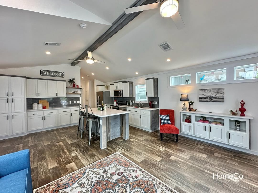 Modern kitchen with white cabinets, island, and barstools. Rustic wood floors, decorative rug, and cozy red chair add warmth. Ceiling beams and fans enhance spaciousness.