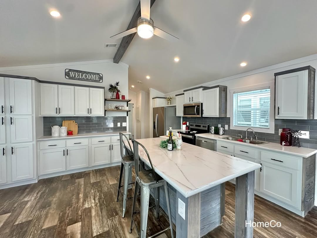 Modern kitchen with white cabinets, gray backsplash, and a large island with two metal chairs. Lit warmly, the space feels inviting and spacious.