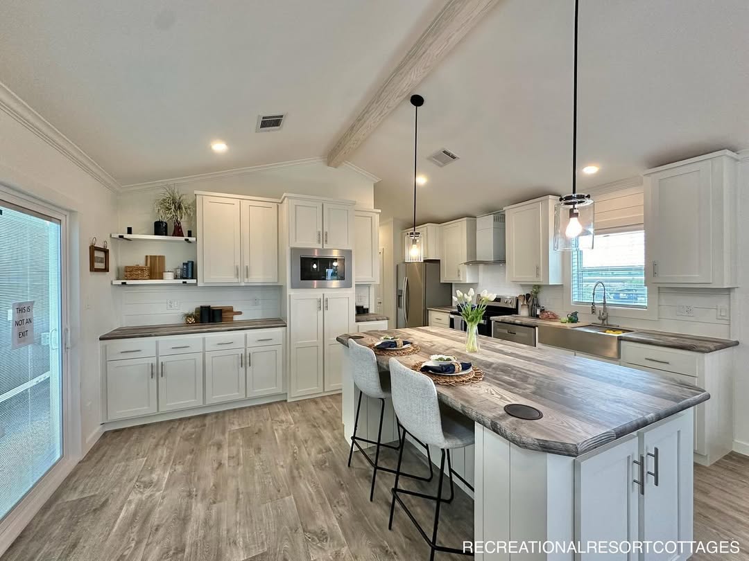 This comprehensive collection of premium tiny homes, park models, and manufactured homes highlights the "Boujee XL" aesthetic, blending modern farmhouse charm with rustic luxury. Exterior architectural styles feature crisp white and cream board-and-batten siding with white metal roofs, black-pillared gabled porches, and elegant black metal railings, as well as a rustic log-cabin style model featuring natural wood siding and a forest green metal roof. Interiors emphasize an upscale lifestyle with vaulted ceilings, exposed wood beams, and white shiplap walls. Gourmet kitchen designs range from rustic wood cabinetry with stone-look backsplashes to sophisticated modern spaces with white or grey cabinetry, marble-look countertops, stainless steel farmhouse sinks, and distressed wood islands. Functional luxury is integrated throughout open-concept living areas, featuring wide-plank wood-look flooring, industrial-style ceiling fans, and large windows or transom glass that provide abundant natural light. Many models also include integrated lofted sleeping quarters accessed by wooden ladders and modern bathrooms with walk-in tile showers.