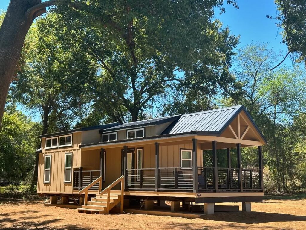 A cozy tan tiny house with a black metal roof sits on a sunny, wooded lot. It has a small porch with railings and steps leading to the entrance.
