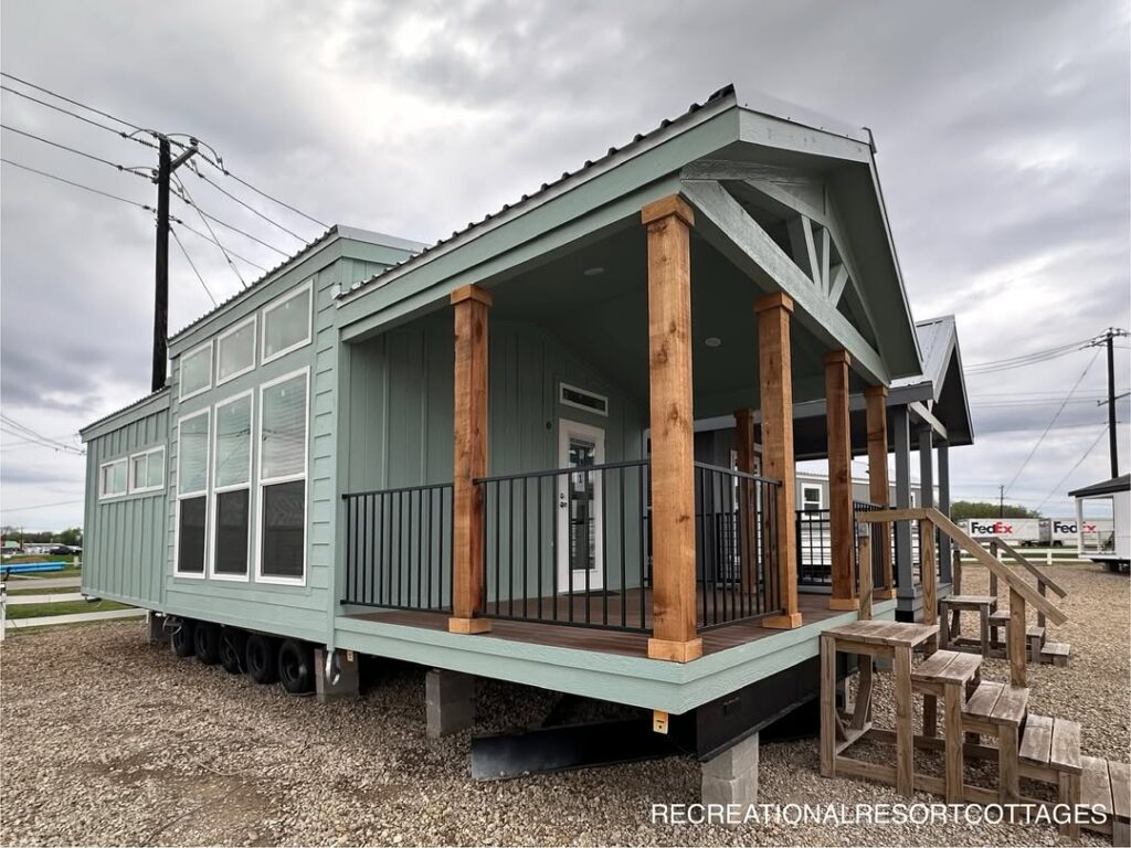 A mint-green tiny house with large windows and a wooden porch stands under a cloudy sky. Wooden steps lead to the porch, creating a cozy and welcoming feel.