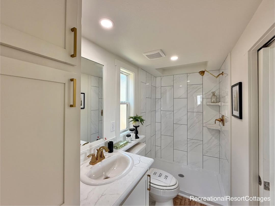 Modern bathroom with white tile walls and gold fixtures. Includes a sink, faucet, and shower. Natural light from a window, potted plant adds greenery.