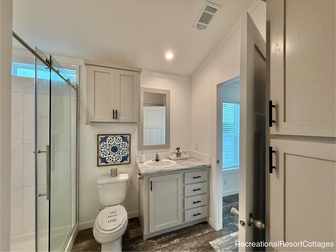 Compact bathroom with glass shower, marble-top vanity, and toilet. Neutral tones with soft lighting and a decorative tile accent. Calm and clean ambiance.