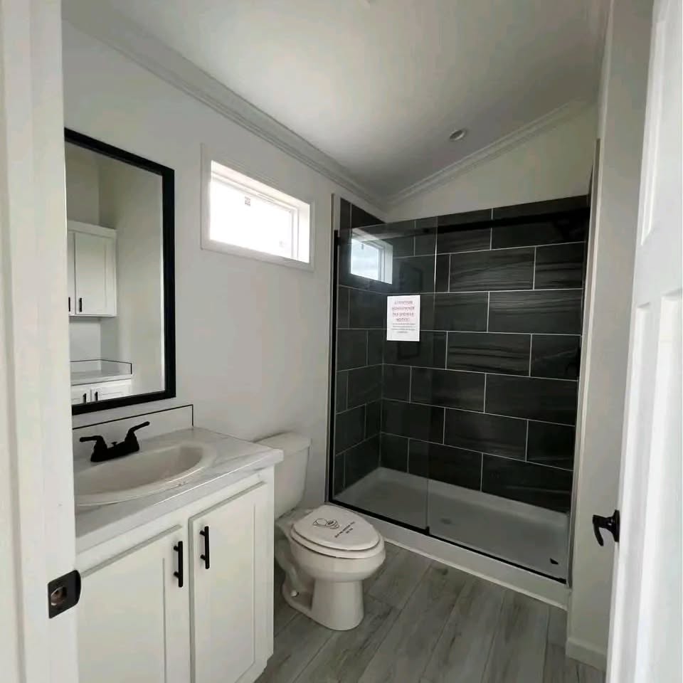 Modern bathroom with a sleek design, featuring a white vanity with a black faucet, a toilet, and a glass-enclosed shower with dark tiles. Bright and clean.