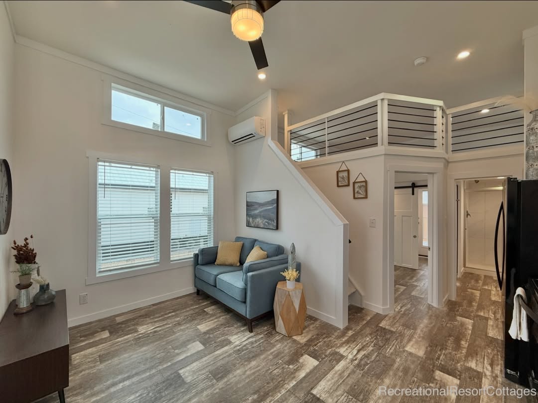 Modern living room with high ceilings and large windows. Features a blue sofa, loft with white railing, wooden decor, and sleek wood flooring. Bright and airy atmosphere.