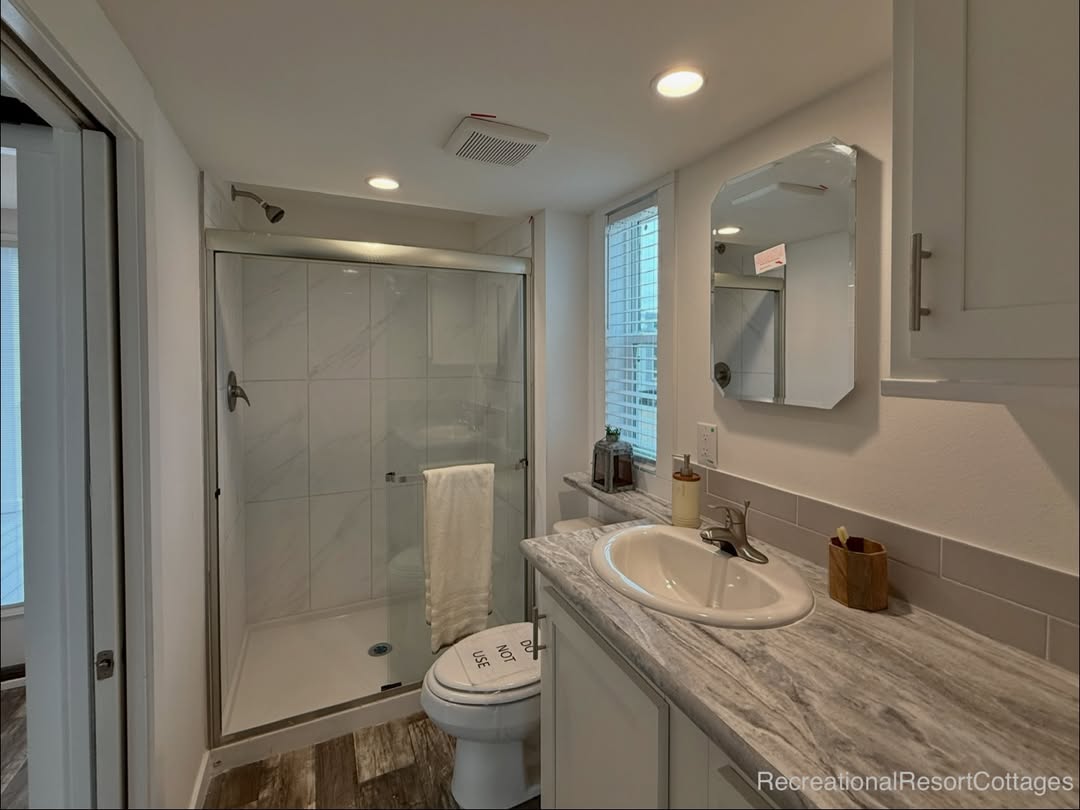 Compact bathroom with cozy lighting, features a glass shower, marble sink countertop, and a window with blinds. Modern, clean, and inviting.