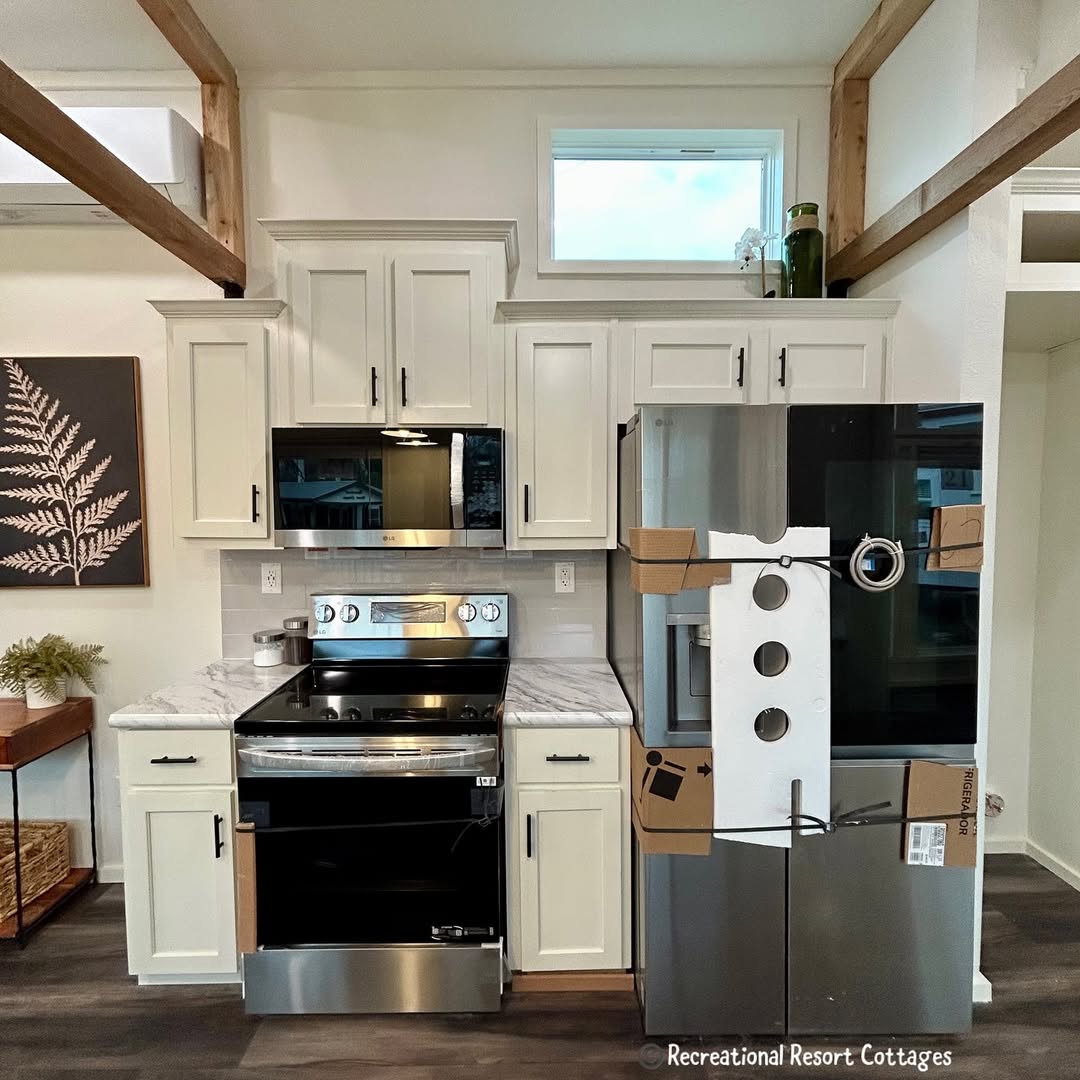 Modern kitchen with white cabinets, stainless steel oven, and marble countertops. A wrapped fridge is on the right. A fern artwork hangs on the left wall. Bright and tidy atmosphere.