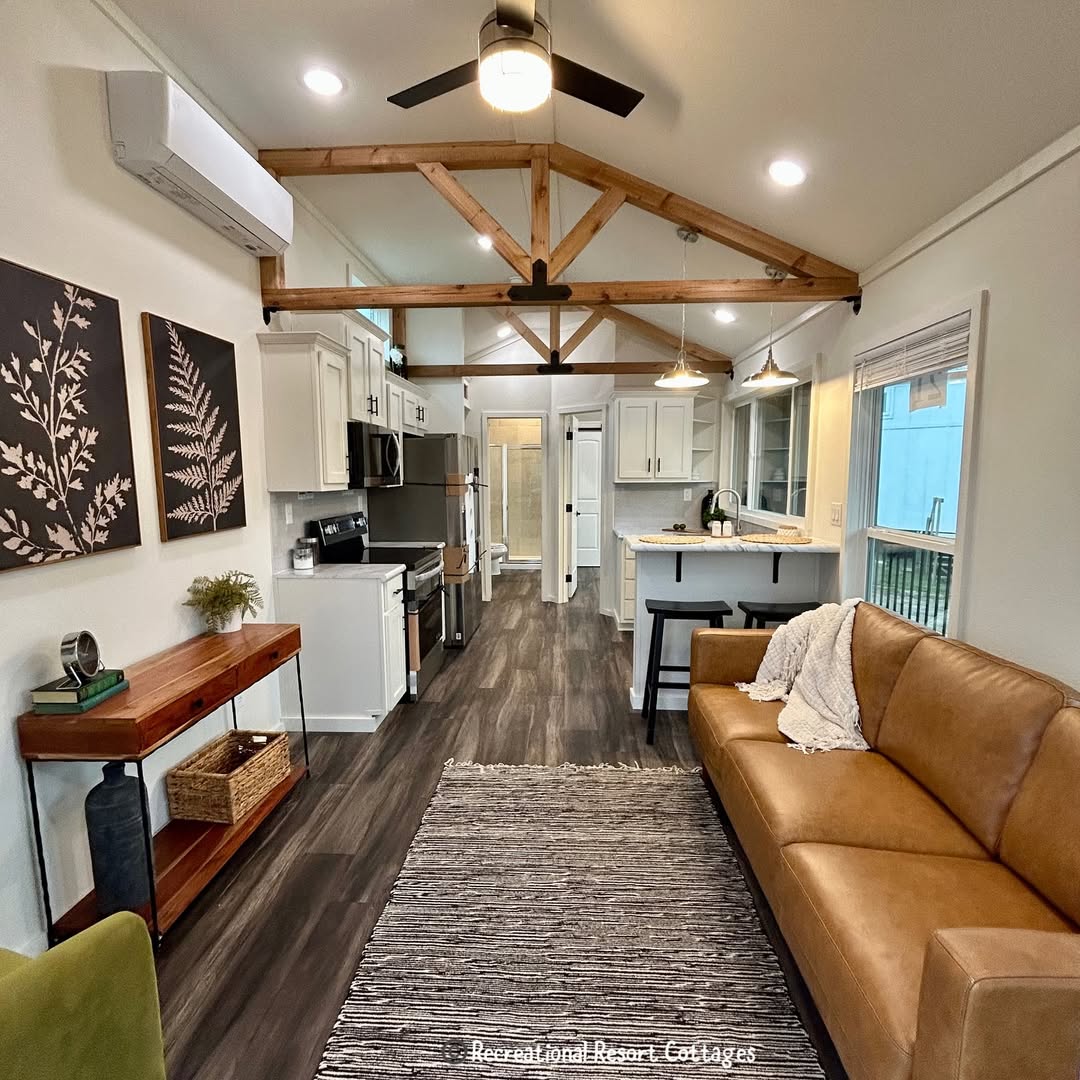Modern, cozy living area with exposed beams, brown leather couch, and kitchen. Warm tones, plants, and fern artwork on a white wall enhance comfort.