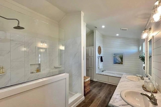 Bright, modern bathroom with a glass shower, dual marble sinks, and wood floor. Artwork and plants add warmth. Cozy, serene ambiance.