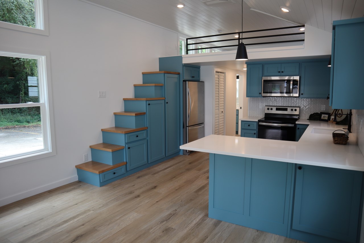 Modern kitchen in a tiny home with teal cabinets, wood accents, and stainless steel appliances. A compact staircase leads to an open loft, creating a cozy feel.