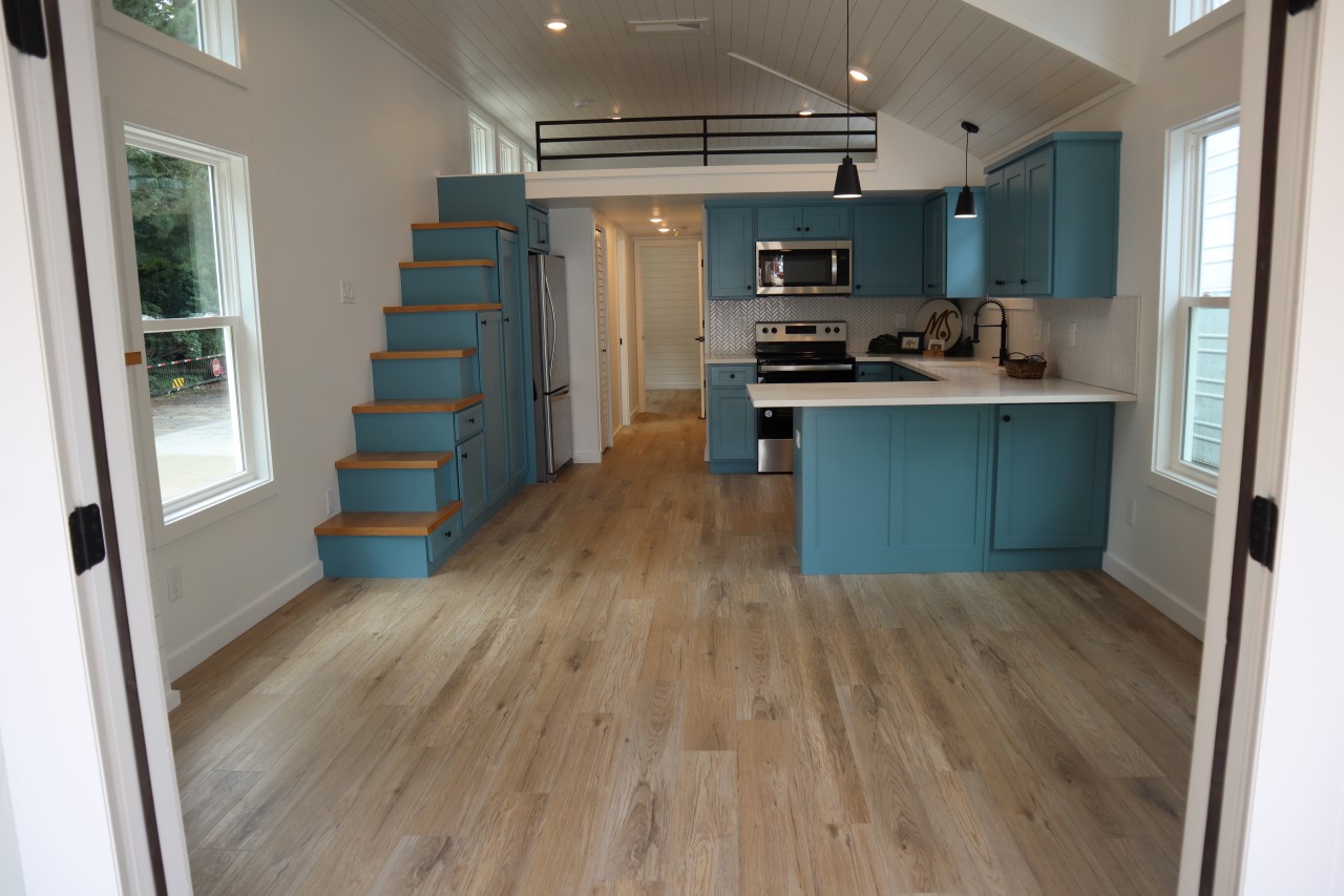 Modern tiny home interior with light wood floors, turquoise cabinets, and white walls. Features a compact kitchen, loft area, and wooden stairs.
