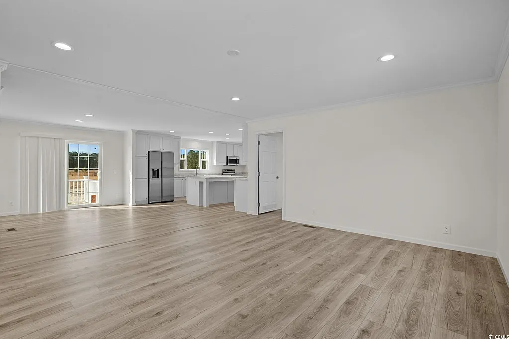 Spacious, empty living room with light wood flooring, cream walls, and recessed lighting. Adjacent modern kitchen features white cabinetry and stainless steel appliances. Bright natural light from sliding glass doors enhances an open, airy feel.