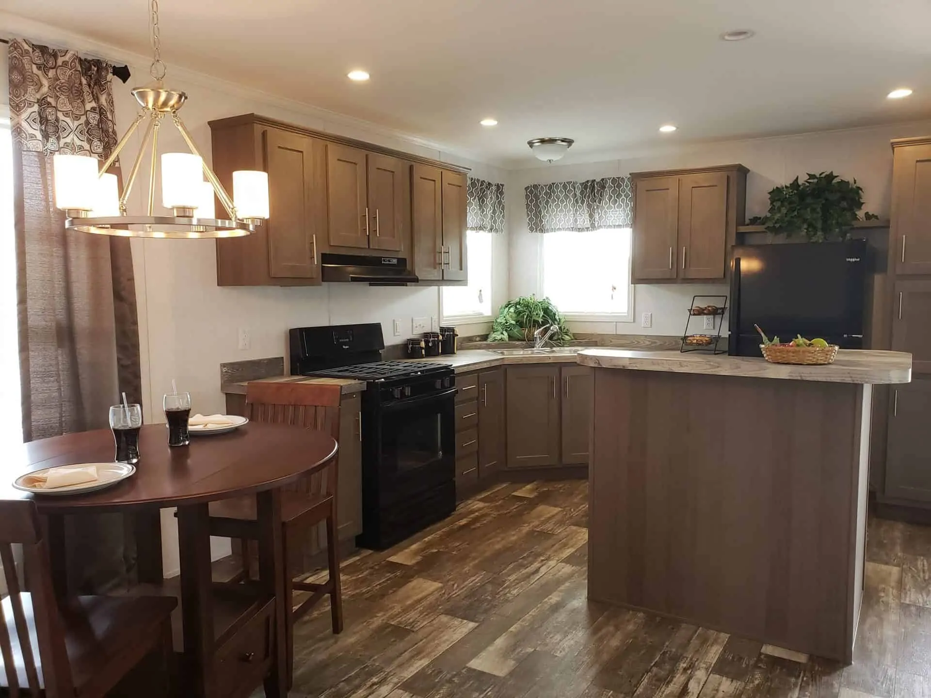 Luxury residential showcase featuring a diverse collection of modern manufactured homes, tiny homes, and park models, including the expansive "The Boujee XL" aesthetic. Exterior Architectural Styles: Highlights include classic white board-and-batten farmhouses with grand pillared front porches and black metal railings, as well as a striking dark charcoal park model with a copper-tone metal roof, natural wood accents, and a wrap-around elevated deck. Designs range from single-level double-wides to compact tiny homes with high-pitched gabled roofs and integrated porches. Interior Living & Design: Open-concept interiors are defined by high-fidelity finishes, including vaulted ceilings with rustic dark wood beams, white shiplap accent walls, and integrated electric fireplaces with custom mantels. Living areas offer a mix of modern luxury and cozy functionality, featuring lofted sleeping quarters with black metal balustrades and natural wood-look flooring throughout. Gourmet Kitchens & Dining: The collection showcases various kitchen layouts, from minimalist tiny home kitchens with natural wood cabinetry and black marble-look countertops to expansive gourmet kitchens. High-end features include oversized islands in statement colors like teal and charcoal, farmhouse-style white cabinetry, professional-grade stainless steel appliances, and elegant pendant lighting. Spa-Inspired Bathrooms: Bathrooms emphasize modern luxury with walk-in tile showers featuring black matte hardware, freestanding soaking tubs with marble-look surrounds, and double vanities with LED-mirrored lighting. This gallery represents the pinnacle of "boujee" manufactured living, combining versatile 4-to-5 bedroom layouts with sophisticated, modern farmhouse aesthetics.