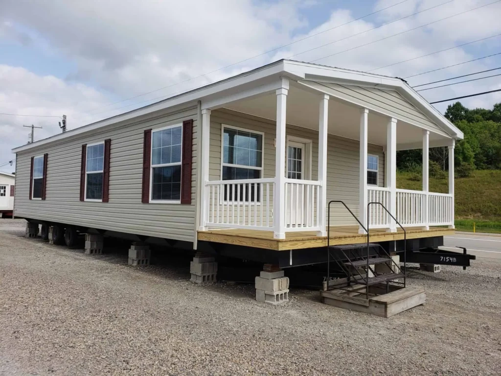 High-end housing showcase featuring a diverse collection of modern manufactured homes and park models, including the expansive "The Boujee XL" style. Exterior Architectural Styles: Highlights include classic white board-and-batten tiny homes with grand pillared front porches and black metal railings. A standout model features a striking dark charcoal exterior with a copper-tone metal roof, natural cedar wood accents, and a large wrap-around wooden deck. Traditional manufactured home designs are also featured, showcasing tan vinyl siding with decorative red shutters and spacious covered porches. Interior Living & Custom Design: Open-concept interiors are defined by high-fidelity finishes, including vaulted ceilings with rustic dark wood beams and white shiplap walls. Living spaces showcase a "modern boujee" style with neutral-toned sofas, integrated electric fireplaces with white mantels, and lofted sleeping quarters. Smaller models feature cozy sitting areas with patterned armchairs and decorative window treatments. Gourmet Kitchens: The collection features diverse kitchen layouts, from rustic wood-cabin style kitchens with dark marble-look countertops and stainless steel refrigerators to high-end modern designs. Premium features include oversized kitchen islands in statement colors like teal and charcoal, white farmhouse cabinetry, recessed lighting, and elegant chandeliers over dining areas with dark wood-look flooring. Spa-Inspired Bathrooms: Bathrooms emphasize modern luxury with walk-in tile showers featuring black matte hardware, marble-look surrounds, and contemporary grey vanities with ample storage. This gallery represents the pinnacle of "boujee" manufactured living, combining versatile floor plans with sophisticated, modern farmhouse aesthetics.