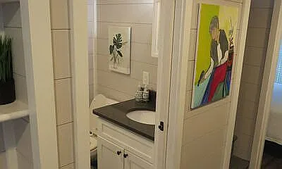 Small bathroom with white tiles, a black countertop, and a round mirror. A colorful painting and a plant add contrast, creating a vibrant, cozy atmosphere.