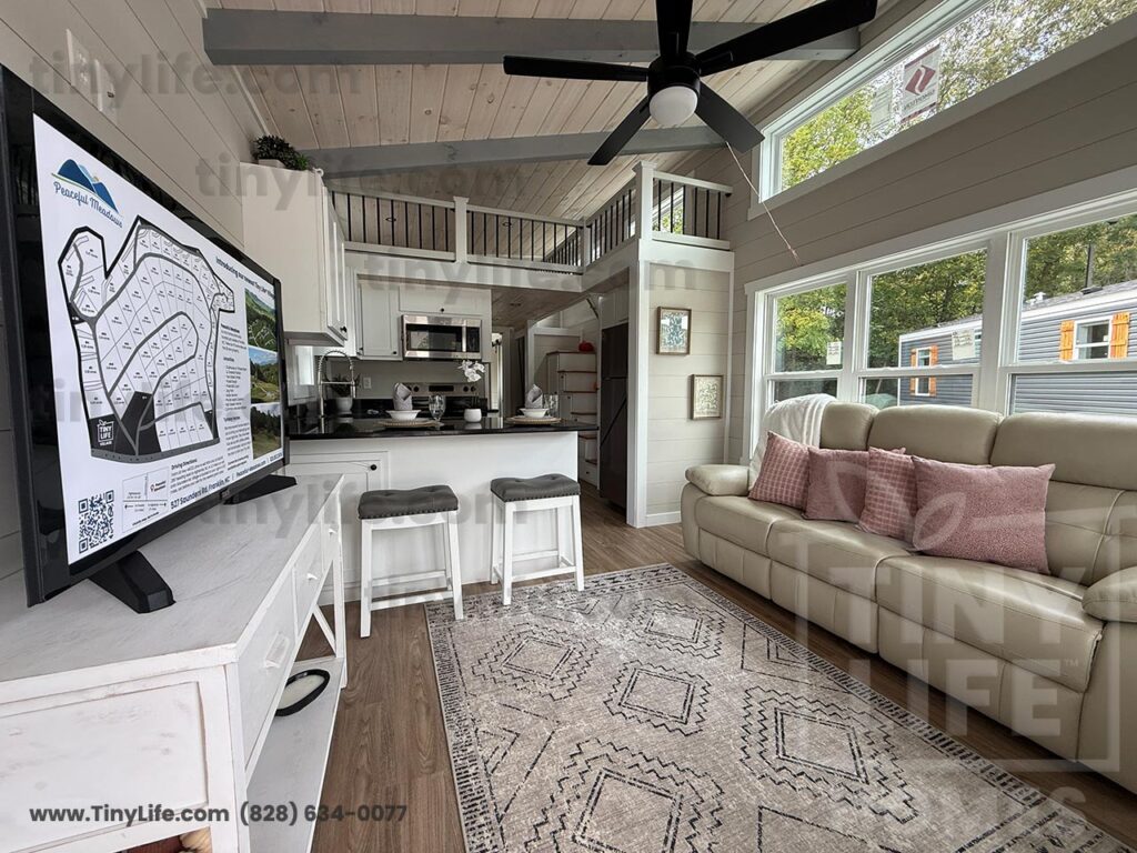 Spacious tiny home living room with large windows, beige sofa, pink cushions, and geometric rug. Ceiling fan above, kitchen and loft visible. Cozy and modern.