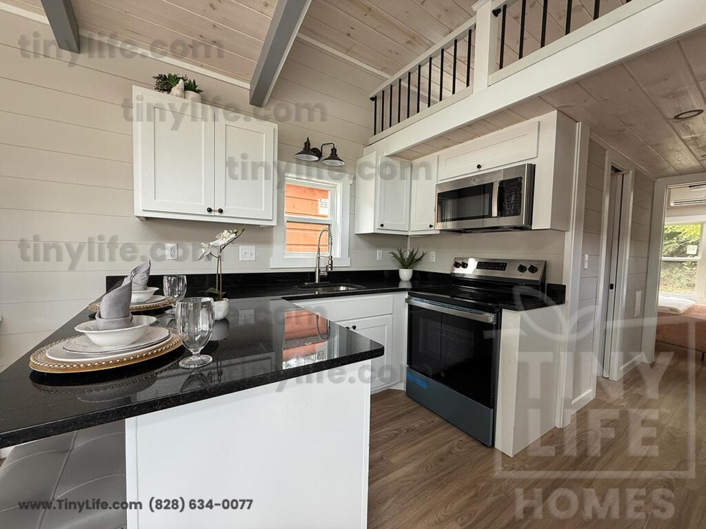 A modern tiny home kitchen with black countertops, white cabinets, and stainless steel appliances. A breakfast bar is set with dishes and glassware.