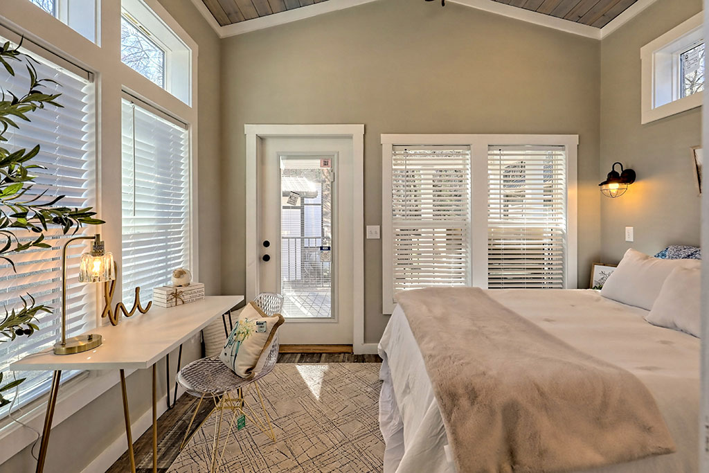 Cozy bedroom with soft lighting features a bed with beige throw, a white desk with a lamp, and a chair. Large windows and a glass door let in natural light.