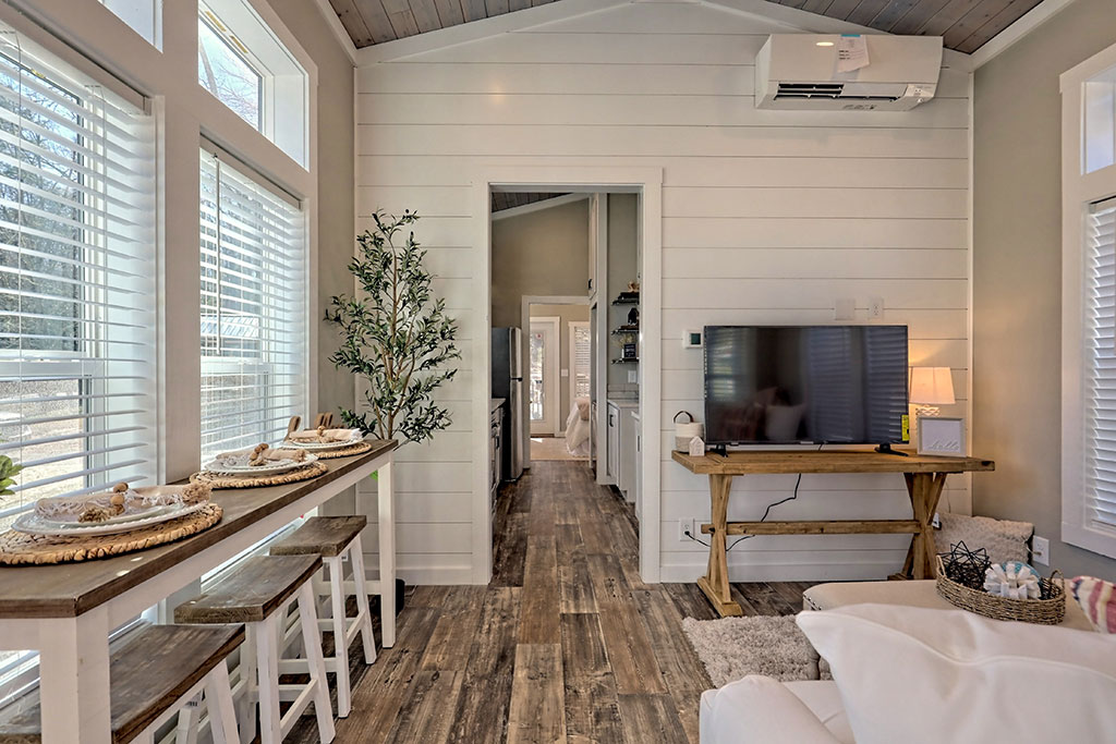Bright living area featuring a cozy wood floor, shiplap walls, large windows with blinds, a small table with stools, and a TV on a rustic stand.