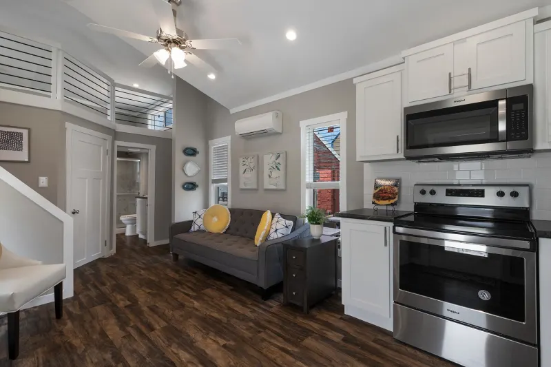 Modern studio apartment with warm wood flooring, a gray sofa with yellow pillows, stainless steel kitchen appliances, and loft area. Bright and airy feel.