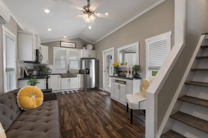 Modern kitchen-living area with white cabinets, stainless steel appliances, and wood floors. A plush gray sofa and cheerful yellow accents add warmth.
