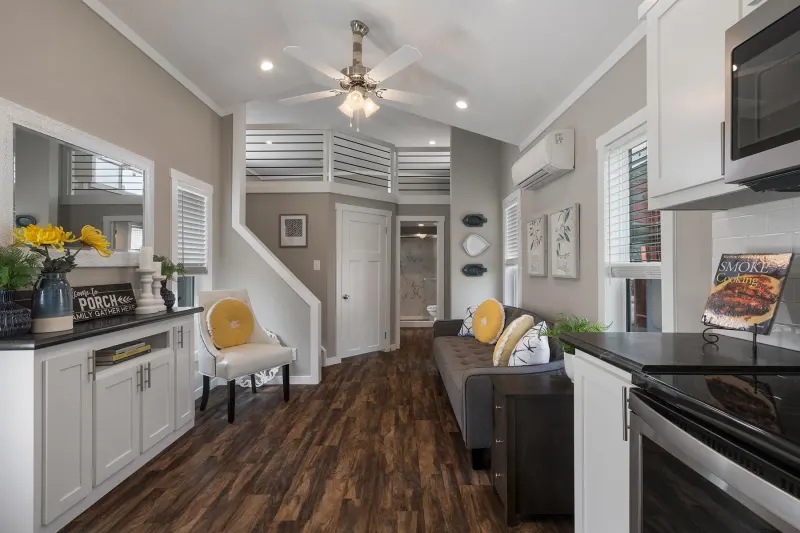 Modern living area with wood flooring, gray walls, and white trim. Features a plush sofa, decorative pillows, indoor plants, and wall art. Bright and inviting.