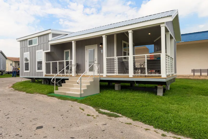 Compact gray tiny house with a blue roof on a green lawn. Features a front porch with chairs, white railing, and stairs leading to the entrance.