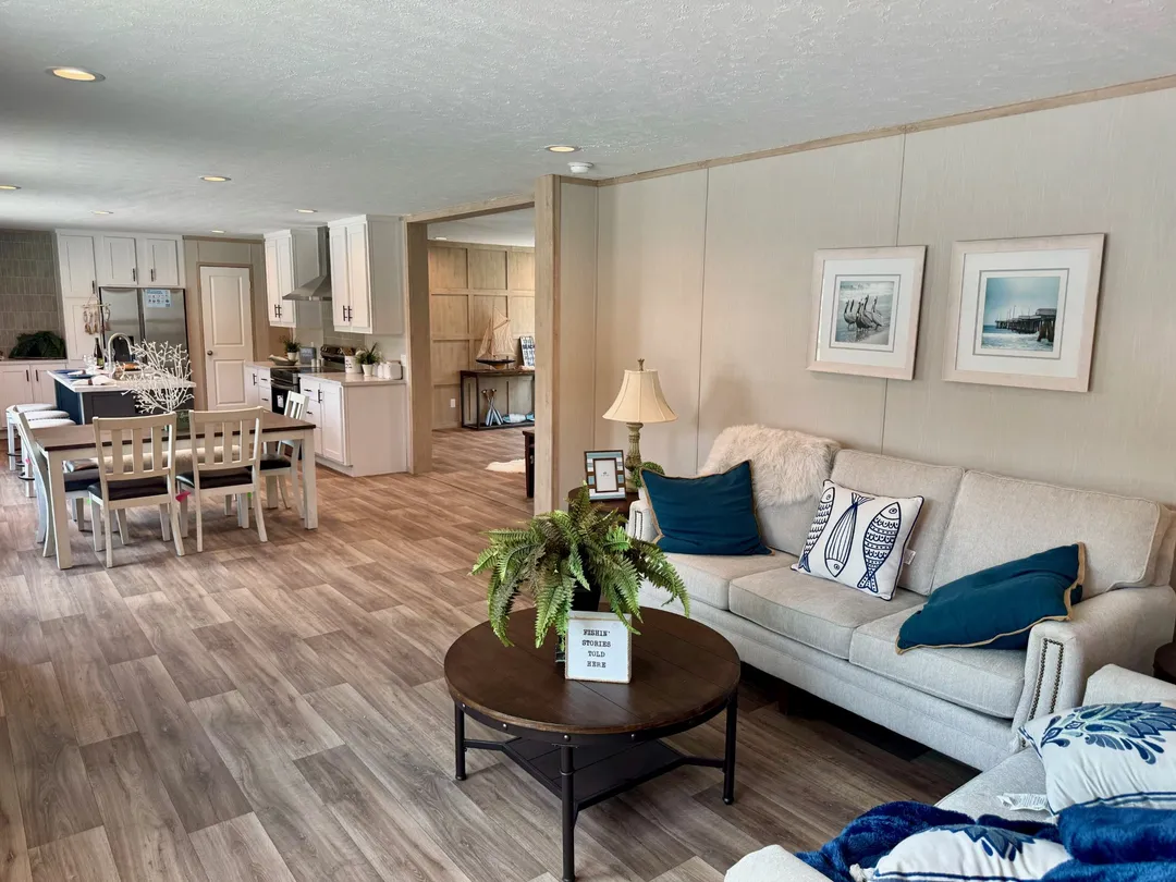 Modern open-plan living room with neutral tones and wooden flooring. Features a beige sofa with blue cushions, a round coffee table, and a dining area. Cozy and inviting atmosphere.