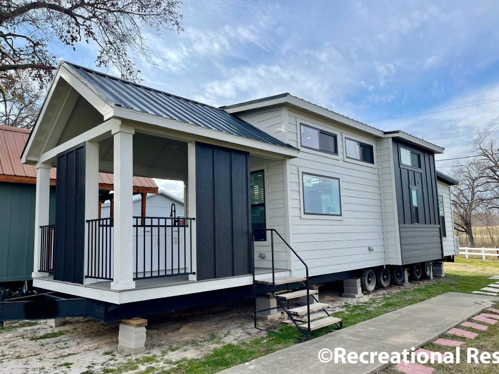 Modern tiny house with wheels, featuring a gray exterior, dark trim, and a small covered porch. Sits on a grassy area under a cloudy sky.