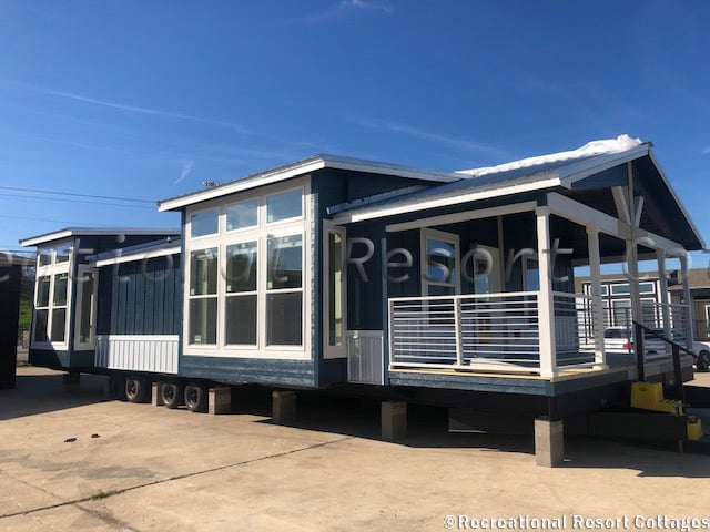 Modern blue tiny home on wheels with large windows and a covered porch sits on a sunny lot, conveying a sense of mobility and contemporary living.