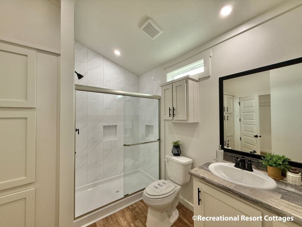 Modern bathroom with a sleek design. Features a glass shower with white marble tile, a vanity with a black-framed mirror, and decor with green plants.